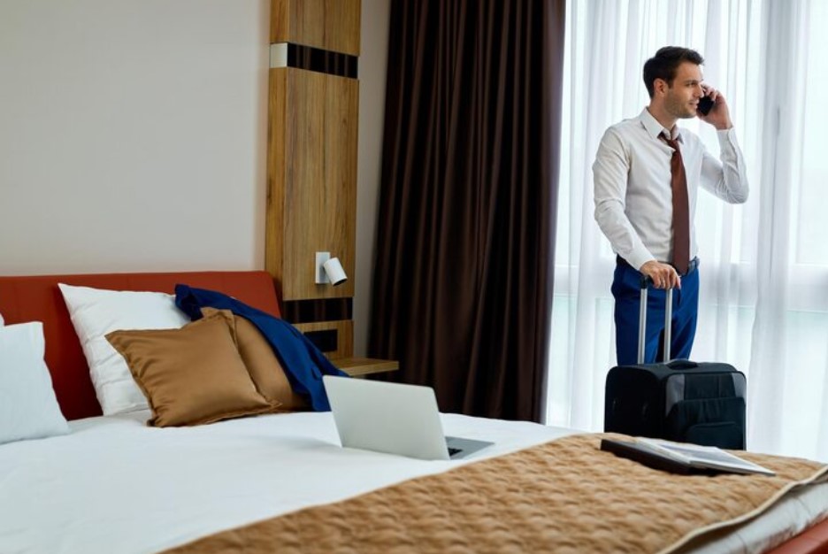 Smiling businessman communicating on mobile phone while standing with a travel bag in hotel room