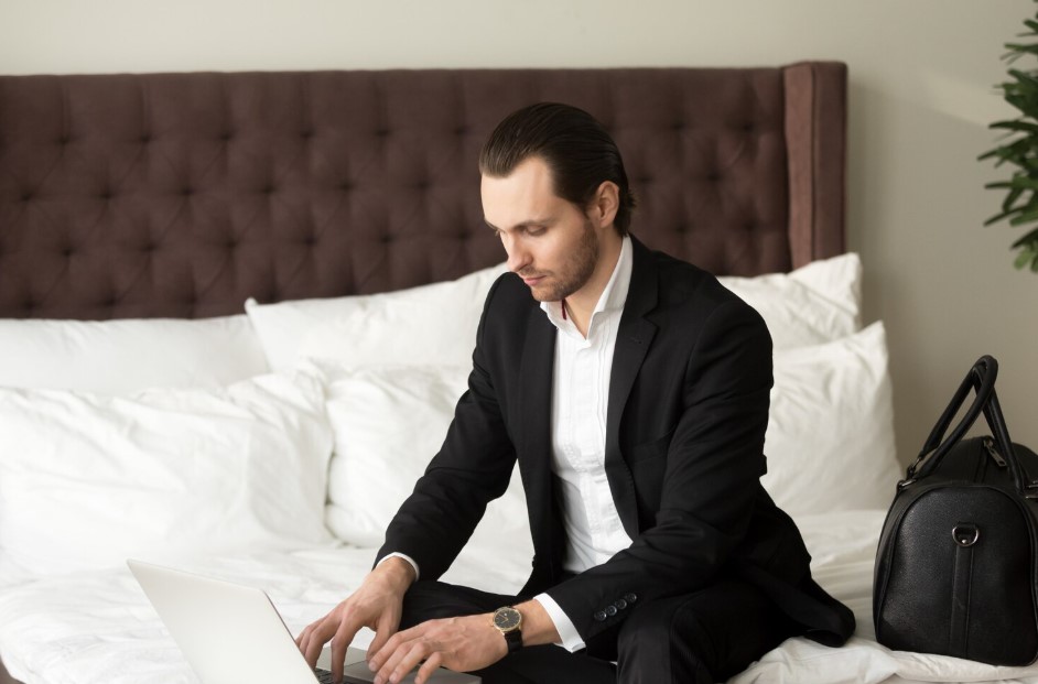 Young businessman sitting on bed working on laptop in hotel.