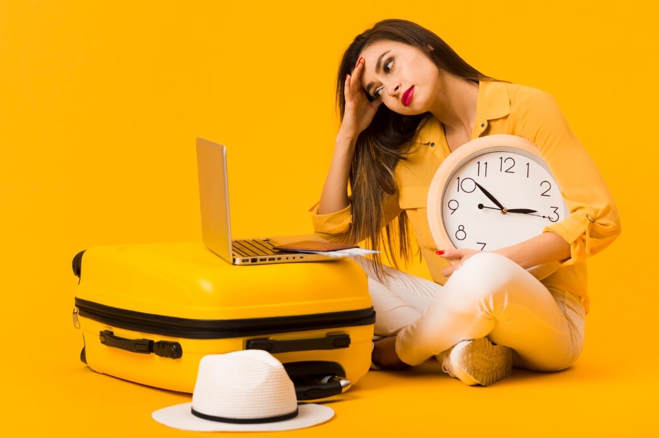 Frustrated woman holding clock and looking at laptop on top of luggage