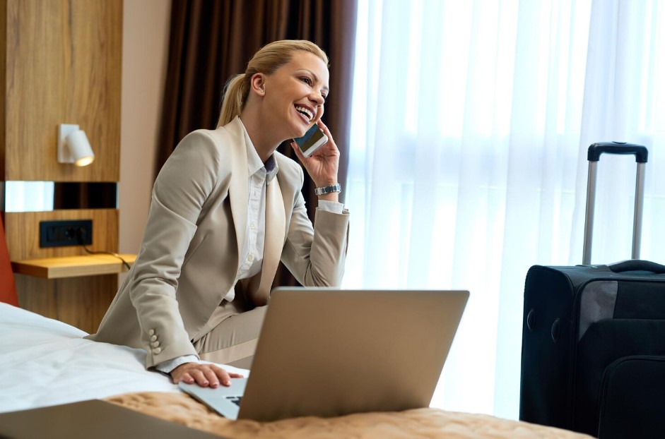 Happy businesswoman sitting on the bed in hotel room and talking on mobile phone